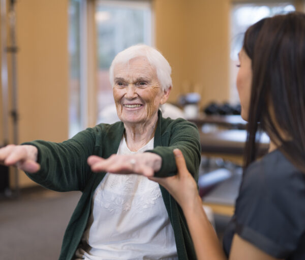 A female occupational therapist of Asian descent works with a cheerful senior female as she does exercises on an exercise ball. She is helping steady her as she holds her arms out.