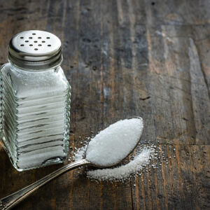 Glass salt shaker shot on rustic wooden table. A spoon with salt is beside the shaker placed directly on the table. The composition is at the left of an horizontal frame leaving useful copy space for text and/or logo at the right. Predominant colors are white and brown. High resolution 42Mp studio digital capture taken with Sony A7rII and Sony FE 90mm f2.8 macro G OSS lens